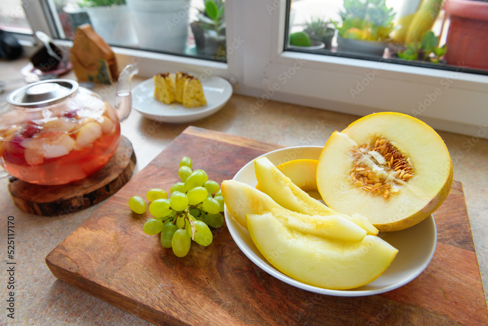 slices of melon, homemade cake on a plate, green grape and a glass teapot with stewed fruit on a kitchen windowsill, concept of fresh fruits and healthy food
