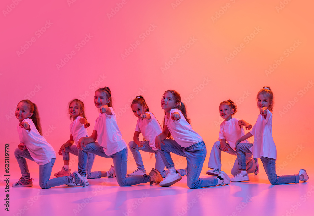 Naklejka premium Happy kids, school age girls in white t-shirts and jeans dancing in choreography class isolated on pink background in yellow neon light.