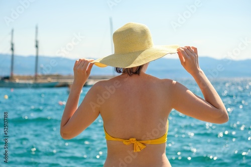 Woman in yellow bikini and straw hat looks at blue sea on warm day. Lady holds headdress with hands against water sparkling in sun close back view © vladim_ka