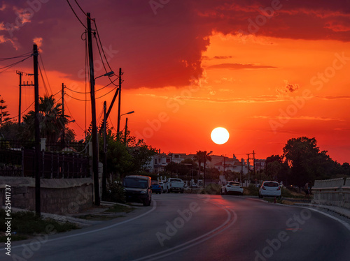 A spectacular sunset illuminates the turn of a paved road. Some cars  electric poles and houses at the entrance of the small town.