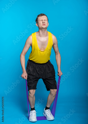 a guy in a yellow T-shirt and black shorts pulled on an elastic band, twisted his face. blue background. hard workout