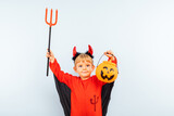 Happy Halloween! Cute little boy in devil halloween costume with trident and pumpkin basket jack-o-lantern on light blue background