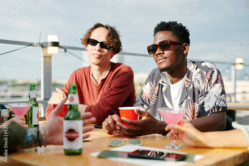Two intercultural guys in sunglasses and casualwear having drinks in outdoor cafe while sitting by table in front of their friends during chat