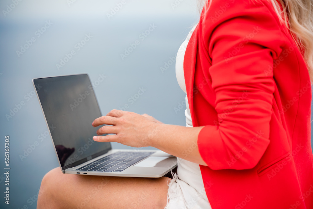 Naklejka premium A woman is typing on a laptop keyboard on a terrace with a beautiful sea view. Close up of a woman's hands writing on a computer. Freelancing, digital nomad, travel and vacation concept.