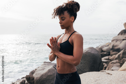 Young woman with folded hands standing at sunset by ocean. Female meditating outdoors with closed eyes.