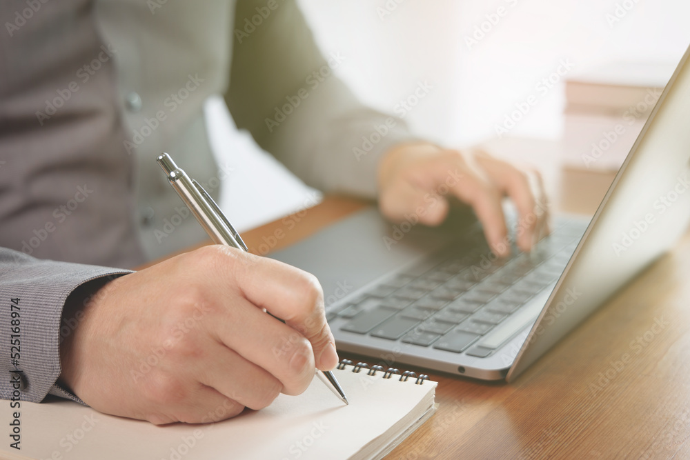 Businessman using laptop for work in the office. He holding pen and writing business information to notebook. Modern communication and work concept. Businessman using laptop for work in the office. He holding pen and writing business information to notebook. Modern communication and work concept.