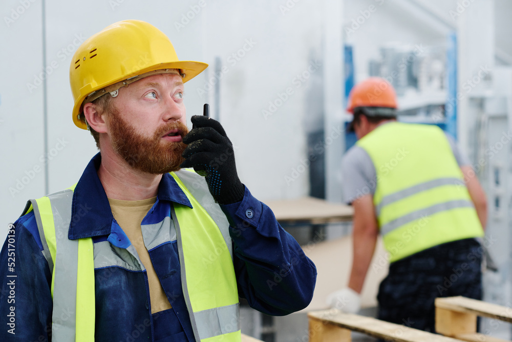 Young confident worker of factory speaking in walkie-talkie while standing in front of camera against colleague in workshop Young confident worker of factory speaking in walkie-talkie while standing in front of camera against colleague in workshop
