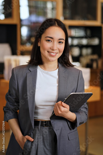 Fotografie Young smiling successful professional leader Asian business woman, female executive manager, saleswoman wearing suit holding digital tablet standing in office looking at camera, vertical portrait