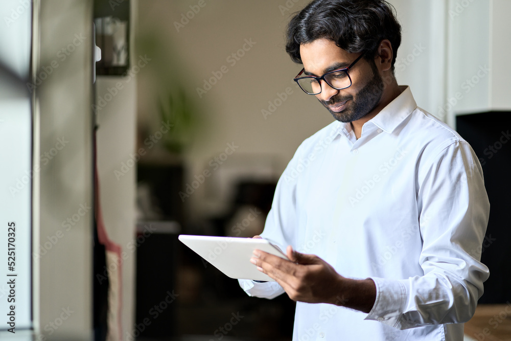 Smiling elegant young indian business man professional company bank manager, arab employee worker, eastern businessman executive standing in office holding using digital tablet and working. Smiling elegant young indian business man professional company bank manager, arab employee worker, eastern businessman executive standing in office holding using digital tablet and working.