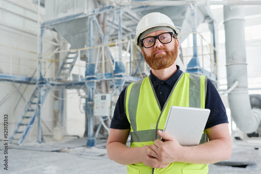 Confident bearded foreman in uniform and safety helmet holding tablet while standing in workshop of modern production factory Confident bearded foreman in uniform and safety helmet holding tablet while standing in workshop of modern production factory
