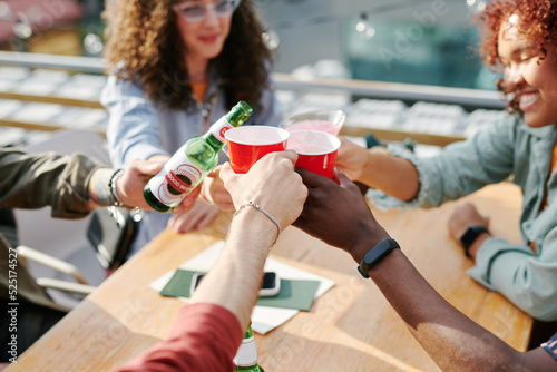 Hands of young interracial friends clinking with drinks ove table in outdoor cafe during rooftop party on sunny summer day