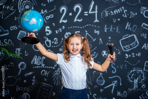 charming mischievous little girl in school uniform, smiling at camera and holding globe and pencils in her hands, having fun during school classes against background black blackboard. Back to school.