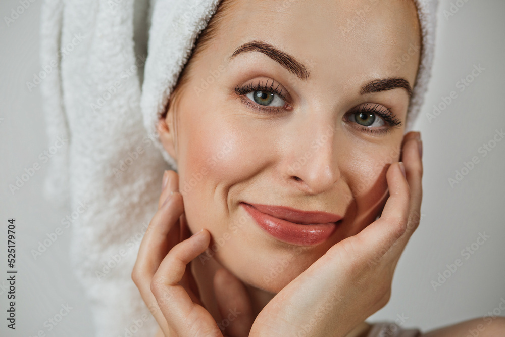 Portrait of smiling caucasian woman with healthy skin in towel posing over grey background. Happy young lady touching her perfect face with hands. Portrait of smiling caucasian woman with healthy skin in towel posing over grey background. Happy young lady touching her perfect face with hands.