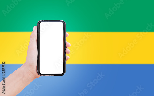 Close-up of male hand holding smartphone with blank on screen, on background of blurred flag of Gabon.