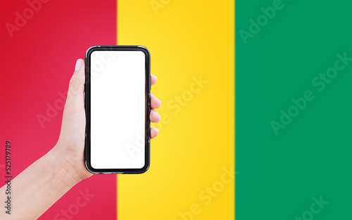 Close-up of male hand holding smartphone with blank on screen, on background of blurred flag of Guinea.