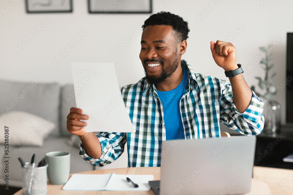 Black Man At Laptop Celebrating Success Holding Paper At Workplace