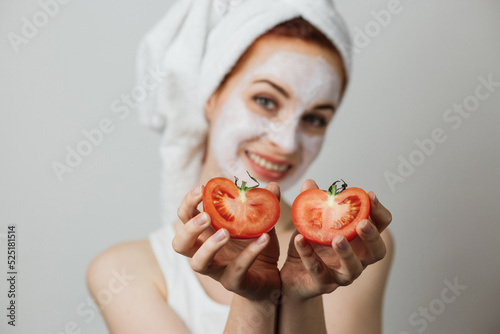 Smiling young woman holding two halves of tomato near her face with white clay mask on face against grey background. Concept of people, skincare and organic cosmetic.
