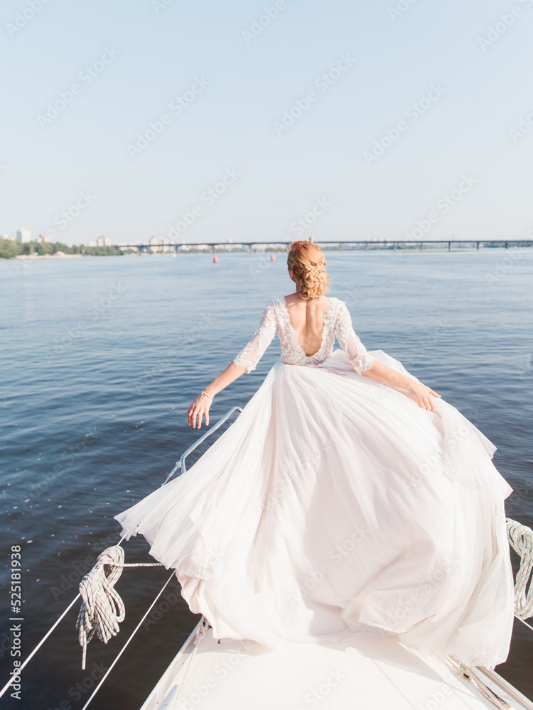 A bride with blond hair in a fluffy white dress with an open back stands on the bow of the yacht with her arms outstretched. The train of the dress is evolving. Ahead is a river landscape and a bridge A bride with blond hair in a fluffy white dress with an open back stands on the bow of the yacht with her arms outstretched. The train of the dress is evolving. Ahead is a river landscape and a bridge