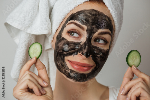 Portrait of caucasian young woman holding slices of cucumber while posing in studio with black clay mask on face. Concept of skin vitaminization and moisturizing. photo