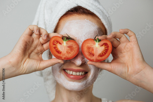 Portrait of caucasian young woman with white cosmetic mask on face and slices of tomato on eyes posing over grey background. Concept of skincare and regular procedure.