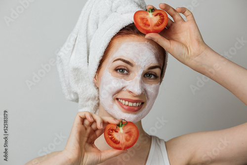 Smiling young woman with white clay mask on face holding fresh tomato against grey background. Concept of aging process and cosmetic procedures.