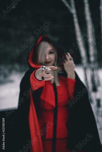 In winter in the forest a witch in a black cloak and red dress stretches her hand forward looking into the camera For halloween a beautiful woman in a witch costume holds a hood looking at the camera
