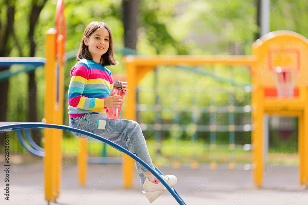 Cute smiling little girl, 5-6 years old, sitting with a plastic bottle of water on the playground