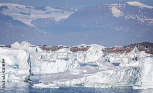 Large icebergs in front of the small community of Saqqaq with towering mountains behind in Disko Bay, Greenland on 18 July 2022 photo