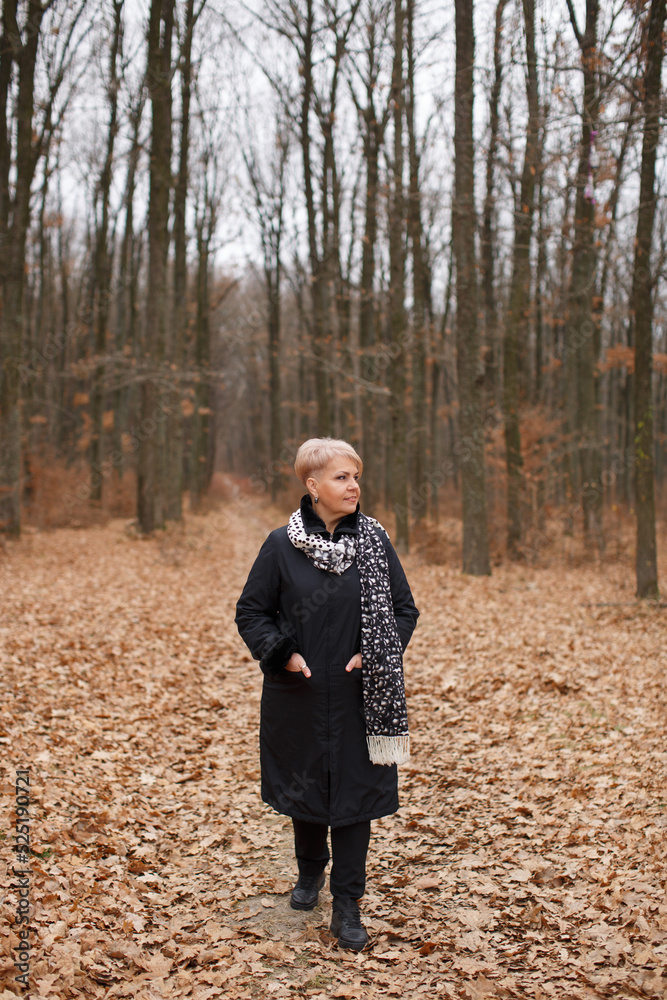 A middle-aged woman with a stylish short haircut in a black coat walks through the autumn forest. A middle-aged woman with a stylish short haircut in a black coat walks through the autumn forest.