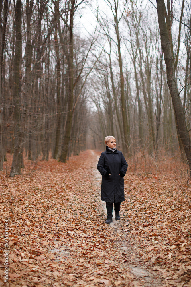 A middle-aged woman with a stylish short haircut in a black coat walks through the autumn forest. A middle-aged woman with a stylish short haircut in a black coat walks through the autumn forest.