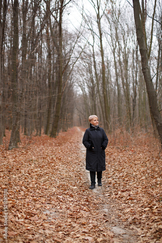 A middle-aged woman with a stylish short haircut in a black coat walks through the autumn forest. 