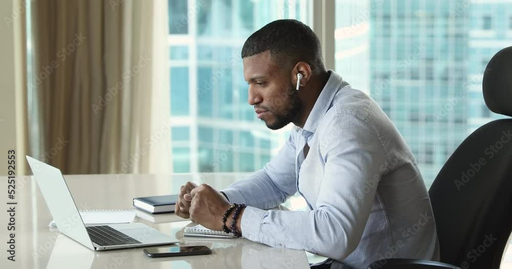 African businessman lead on-line negotiations sit at workplace with laptop, wear wireless earphones take part in distance briefing, solve business work in modern skyscraper office, video call concept African businessman lead on-line negotiations sit at workplace with laptop, wear wireless earphones take part in distance briefing, solve business work in modern skyscraper office, video call concept