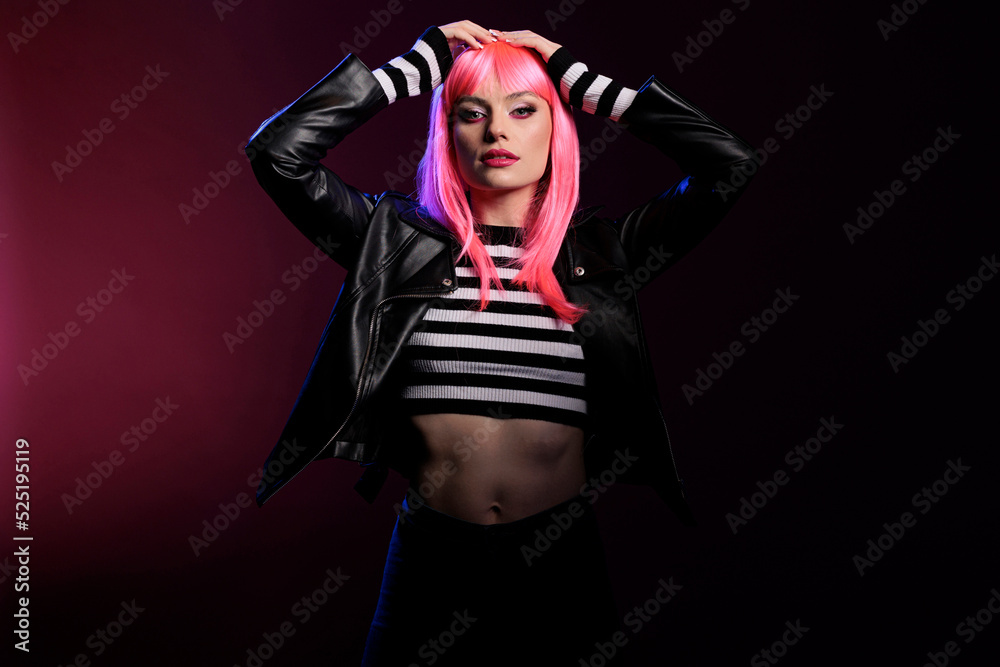 Attractive trendy girl with pink hair wearing leather jacket in studio with dark light, posing over background. Confident woman with stylish rocker clothes and perfect makeup look, fashion style.
