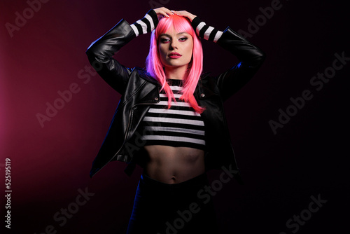 Attractive trendy girl with pink hair wearing leather jacket in studio with dark light, posing over background. Confident woman with stylish rocker clothes and perfect makeup look, fashion style.