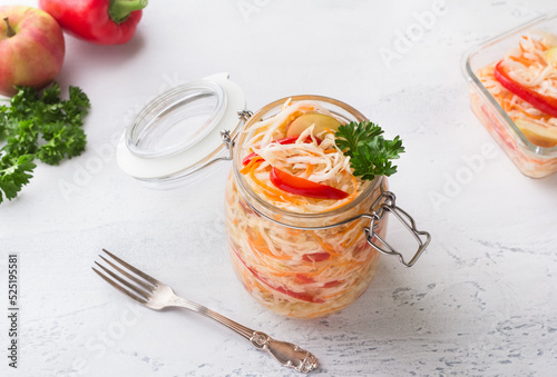 A jar of quick pickled cabbage with carrots, apples and red bell peppers, garnished with parsley on a light blue background. The concept of delicious homemade vegan food