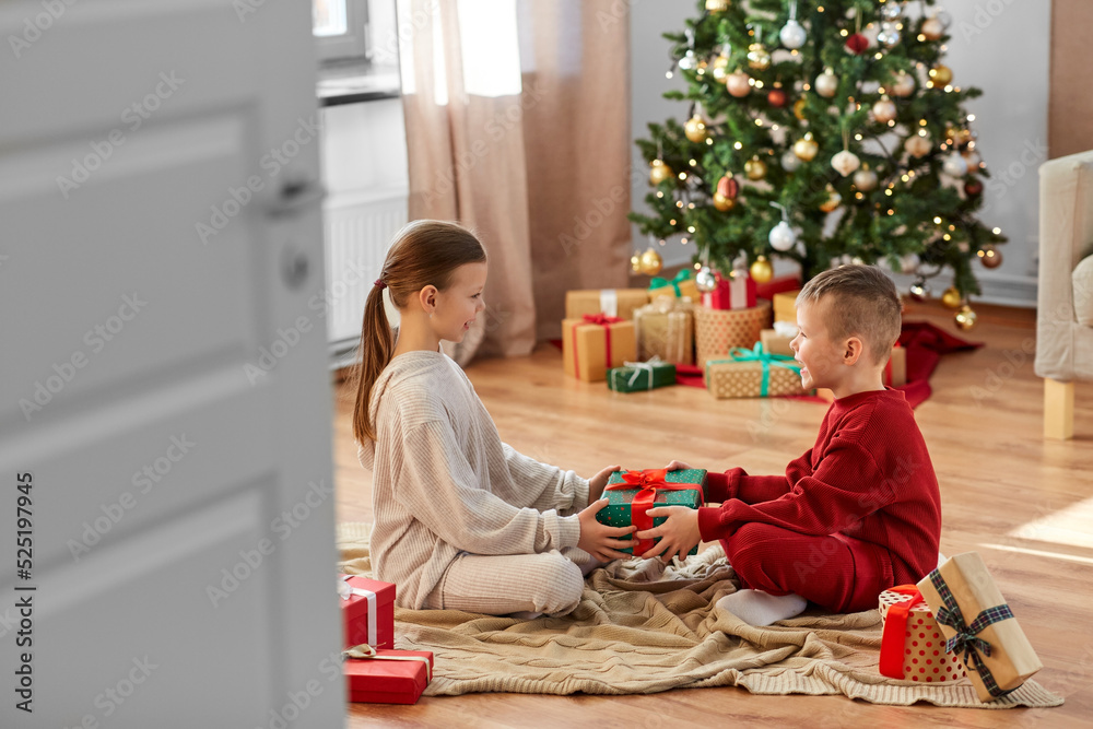 christmas, winter holidays and childhood concept - happy girl and boy in pajamas holding gift box together sitting on floor in front of each other at home
