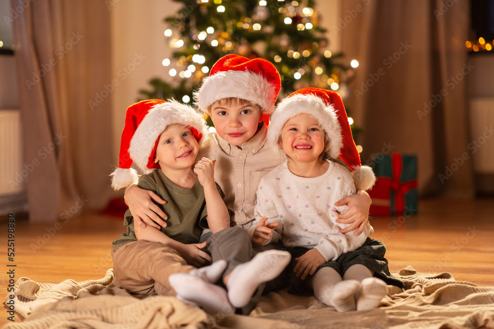 christmas, winter holidays and childhood concept - happy little girl and boys in santa hats hugging on floor at home christmas, winter holidays and childhood concept - happy little girl and boys in santa hats hugging on floor at home