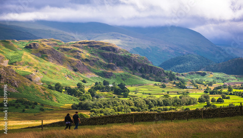 Picturesque view of mountain range in English Lake District at sunset in early fall with farm house and grazing sheep in distance; moody sky in background