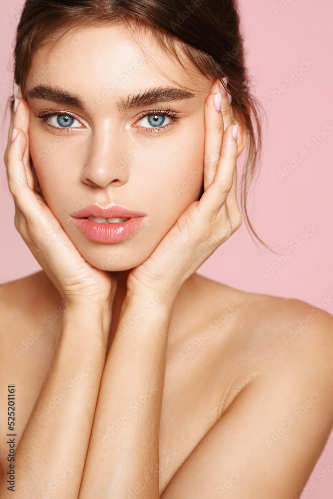 Fototapeta premium Beauty face. Woman with natural makeup and healthy skin portrait. Beautiful girl model touching fresh glowing hydrated facial skin on pink background closeup. Skin care concept