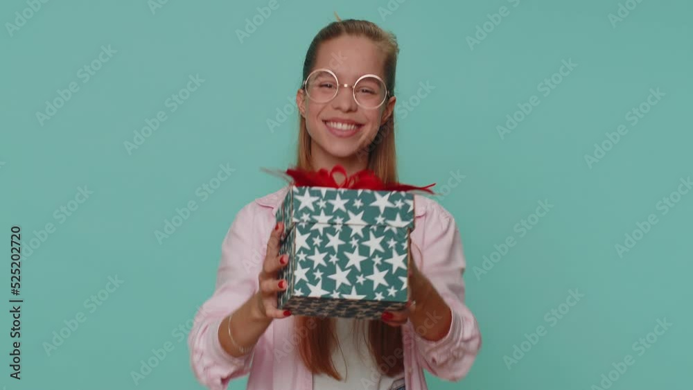 Positive smiling lovely pretty teenager girl presenting birthday gift box stretches out hands, offer wrapped present career bonus, celebrating party. Young child kid on blue studio background indoor
