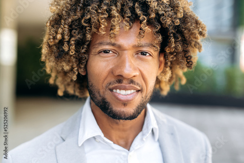 Close-up photo of a handsome charismatic confident curly haired brazilian or hispanic man, successful entrepreneur, standing outdoors, looking at the camera, smiling friendly