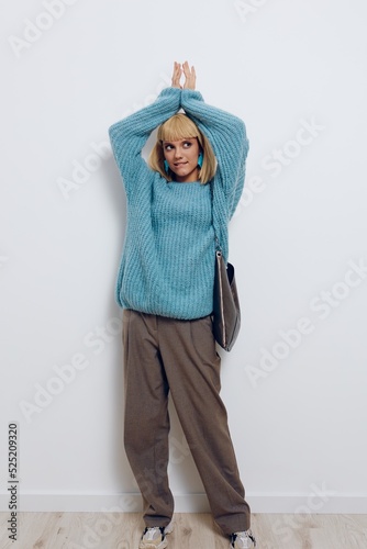 a full-length vertical photo of a beautiful, stylish woman in fashionable earrings and a blue sweater with a large bag and raising her hands up