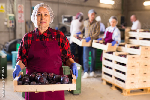 Mature woman in uniform with wooden box full of plums standing in fruit warehouse.