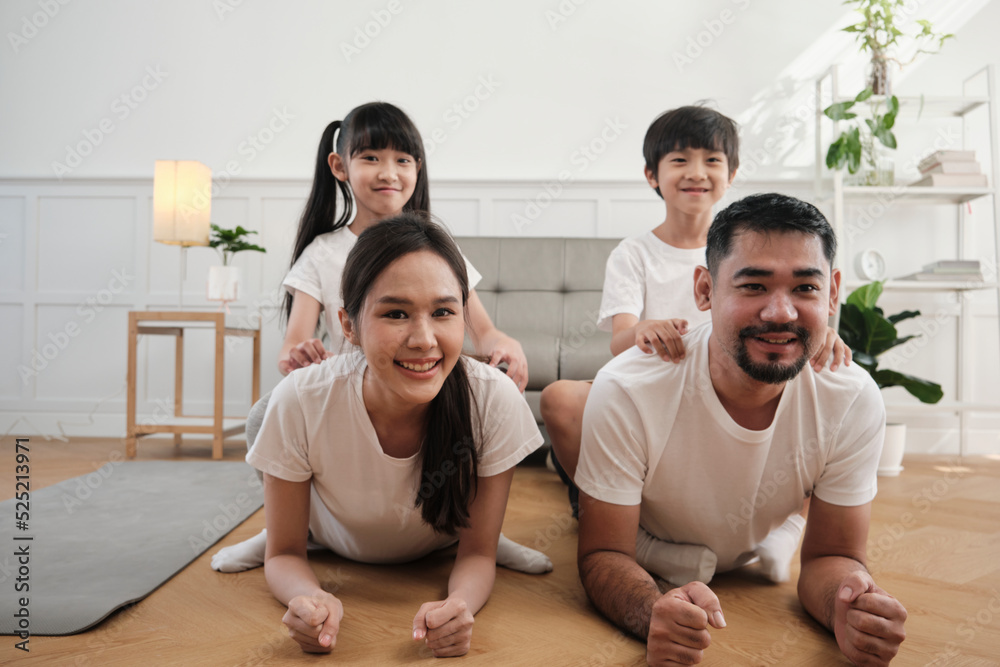 Happy well-being Asian Thai family, children play and tease their parents while yoga fitness training and health exercise together in the white living room, domestic home lifestyle, weekend activity.