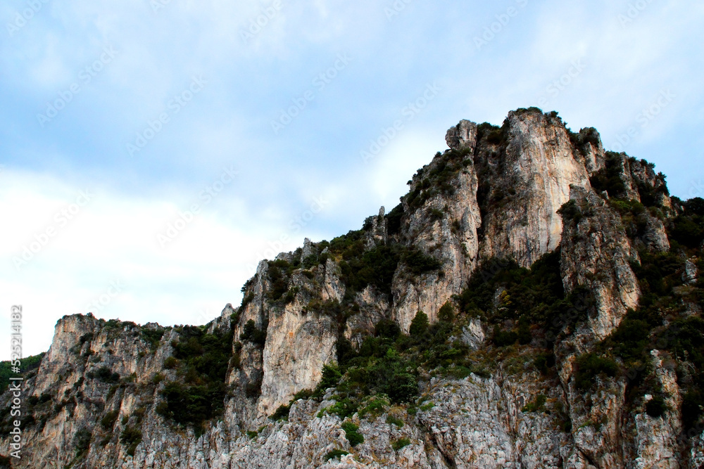 Harsh outlook at a very steep mountain ridge in Amalfi coast punctuated by green vegetation and standing out against a background of hazy azure sky with milky clouds on a summer day Harsh outlook at a very steep mountain ridge in Amalfi coast punctuated by green vegetation and standing out against a background of hazy azure sky with milky clouds on a summer day