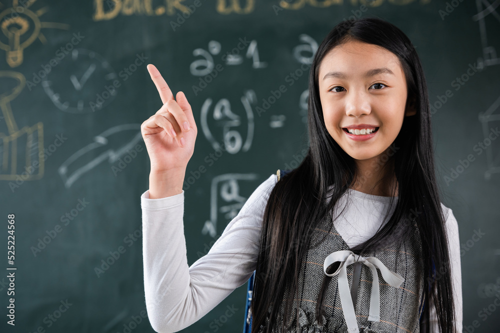 Education Back to school concept. Asian school girl in uniform with backpack on classroom pointing up finger on blackboard, primary child is standing front of class in school thinking of great ideas Education Back to school concept. Asian school girl in uniform with backpack on classroom pointing up finger on blackboard, primary child is standing front of class in school thinking of great ideas
