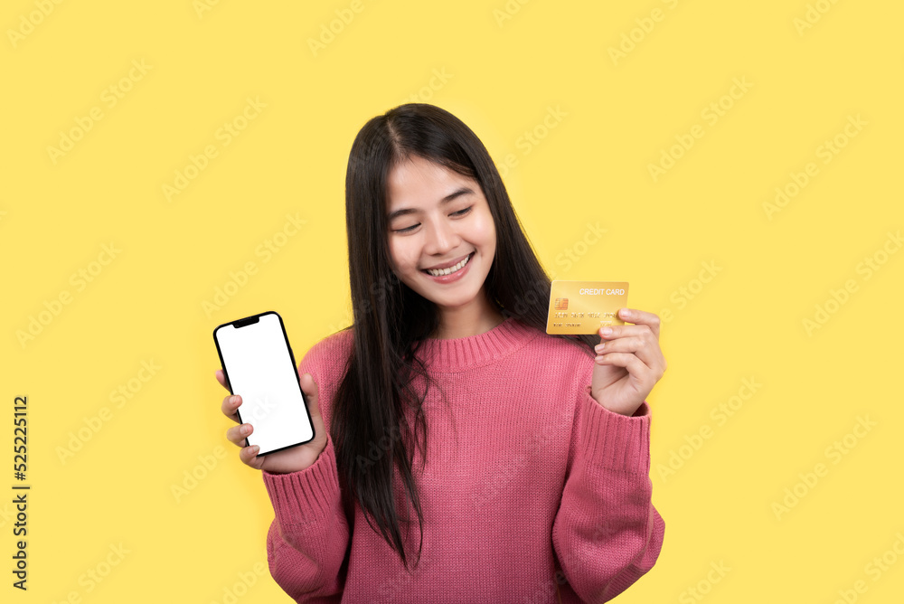 Portrait of a delighted woman standing on a yellow background. holding a cell phone showing a plastic credit card - obrazy, fototapety, plakaty 