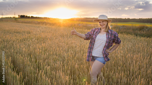 A beautiful middle-aged farmer woman in a straw hat and a plaid shirt stands in a field of golden ripening wheat during the daytime in the sunlight