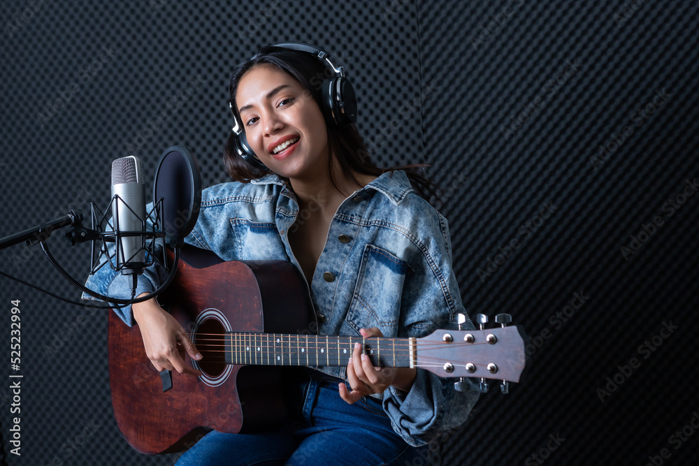 Obraz premium Happy cheerful pretty smiling of portrait a young Asian woman vocalist Wearing Headphones with a Guitar recording a song front of microphone in a professional studio