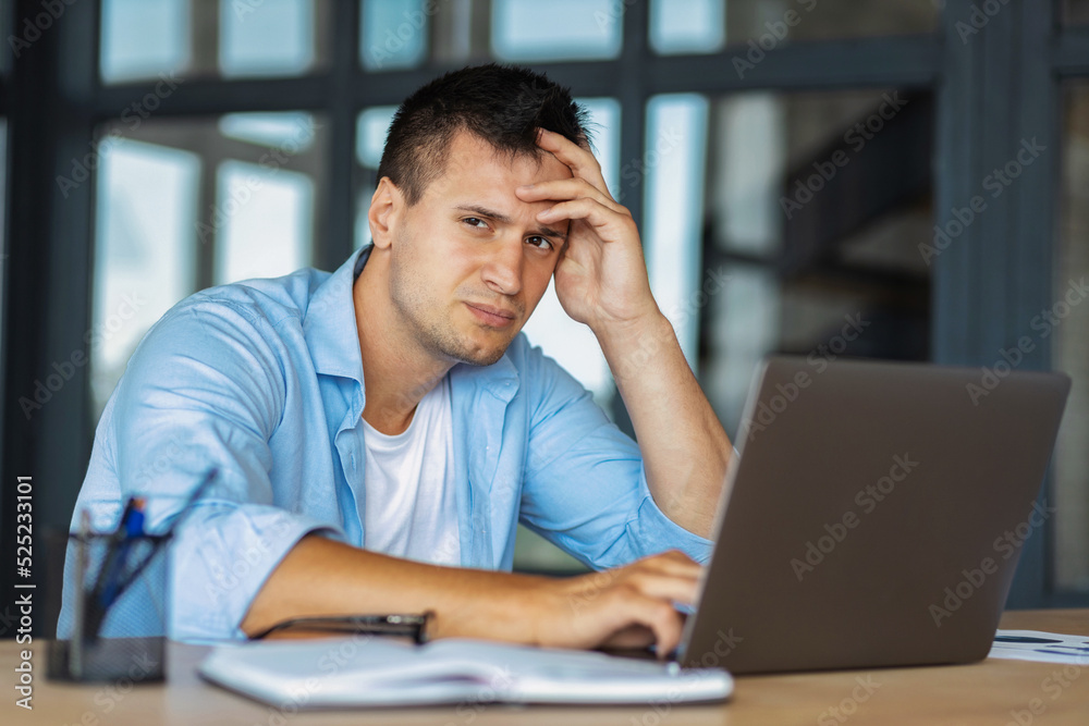 Upset businessman holding head while sitting at workplace in office with laptop. Young business person, got bad news on email, experiences negative emotions. Bankruptcy concept Upset businessman holding head while sitting at workplace in office with laptop. Young business person, got bad news on email, experiences negative emotions. Bankruptcy concept
